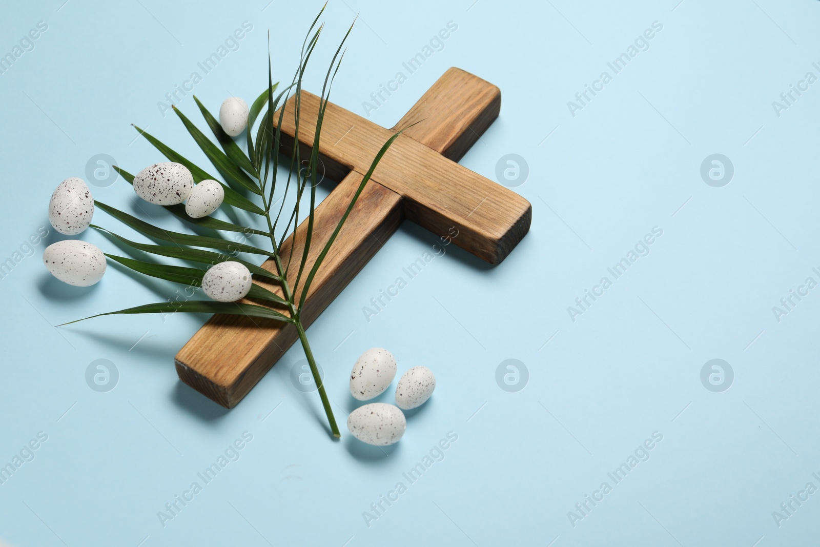 Photo of Wooden cross, painted Easter eggs and palm leaf on light blue background, space for text