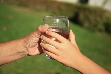 Photo of Child giving glass of water to elderly woman outdoors on sunny day, closeup