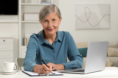 Beautiful senior woman taking notes near laptop at white table indoors