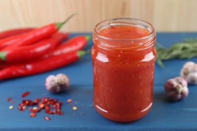 Photo of Spicy chili sauce in jar, garlic, peppers and rosemary on blue wooden table, closeup