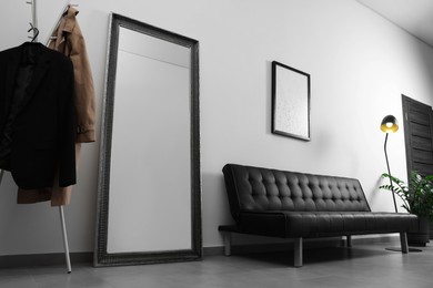 Photo of Coat rack, stylish leather sofa and large mirror near white wall in hallway. Interior design