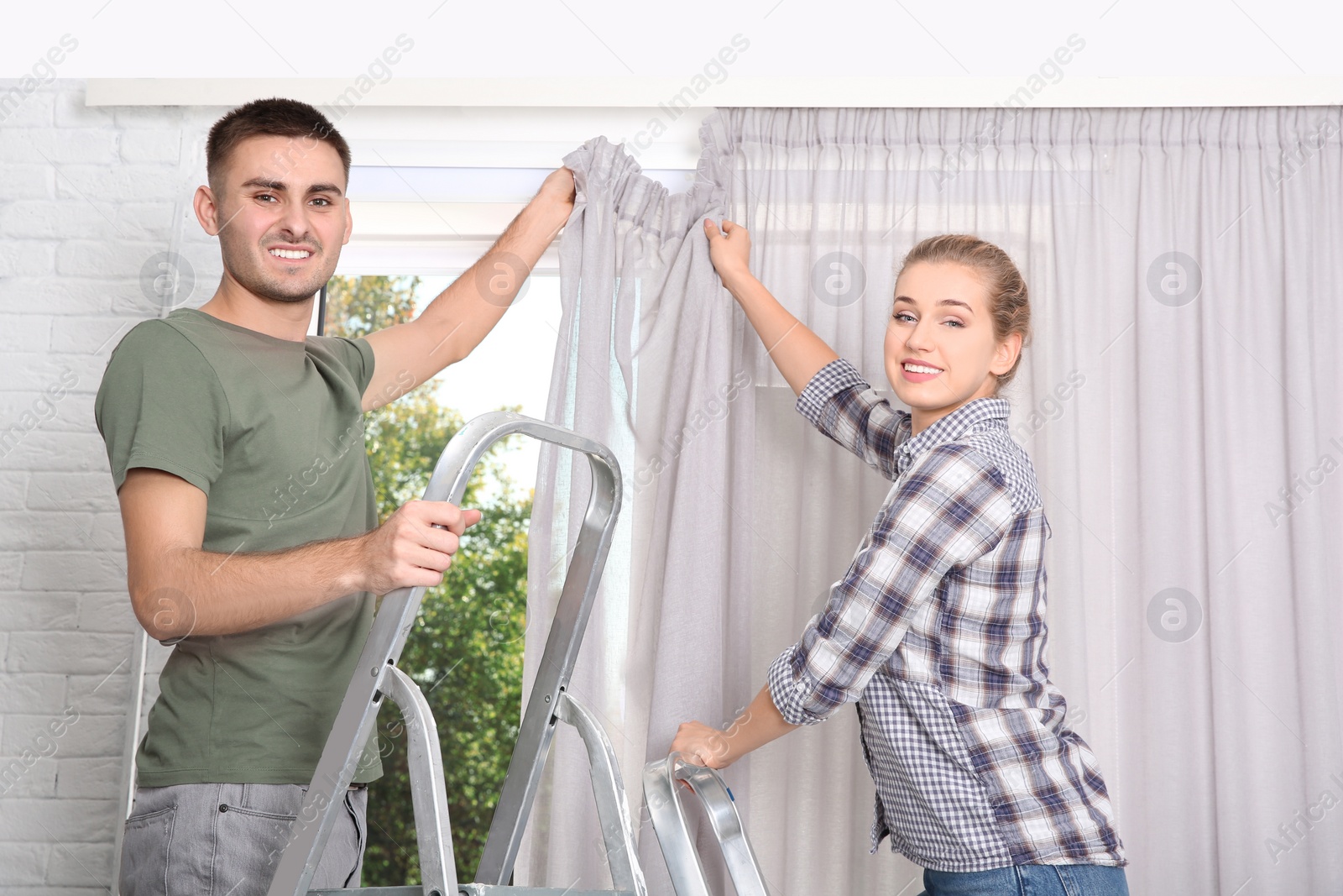 Photo of Young couple hanging window curtain indoors. Interior decor element