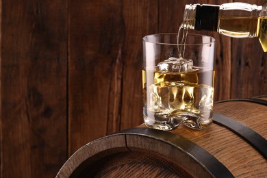 Pouring whiskey from bottle into glass on barrel against wooden background, closeup. Space for text