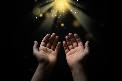 Christian man stretching hands towards holy light in darkness, closeup. Prayer and belief