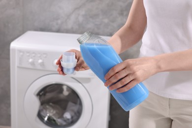 Photo of Woman pouring fabric softener from bottle into cap near washing machine indoors, closeup