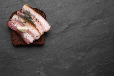 Photo of Tasty pork fatback with rye bread slice, garlic and dill on black table, top view. Space for text