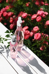 Photo of Bottles of facial toner with essential oil on table near rose bushes. Space for text