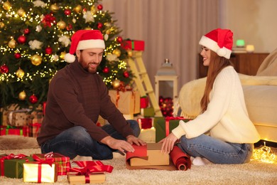 Happy couple in Santa hats decorating Christmas gift with wrapping paper at home