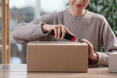 Photo of Young woman using utility knife to open parcel at wooden table indoors, closeup