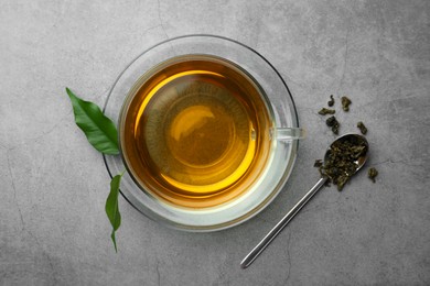 Photo of Fresh green tea in glass cup and spoon with dried leaves on grey table, flat lay