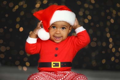Image of Cute little African American baby wearing Santa hat against blurred lights on dark background. Christmas celebration