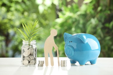 Pension savings. Figure of senior man, piggy bank, jar with coins and twig on white table against blurred green background