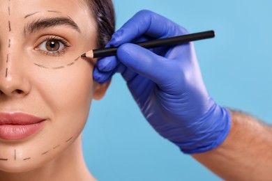 Doctor drawing marks on woman's face for cosmetic surgery operation against light blue background, closeup
