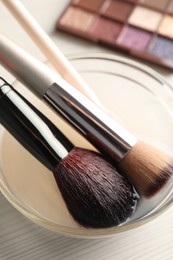 Photo of Cleaning makeup brushes in bowl with special liquid on white wooden table, closeup