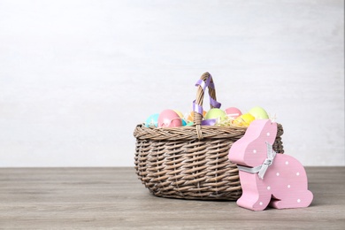 Photo of Cute wooden bunny and wicker basket with Easter eggs on table against light background, space for text