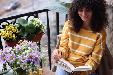 Photo of Young woman reading book at table with beautiful flowers on balcony
