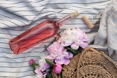 Photo of Bottle of rose wine near straw bag with beautiful peonies on light blanket, flat lay