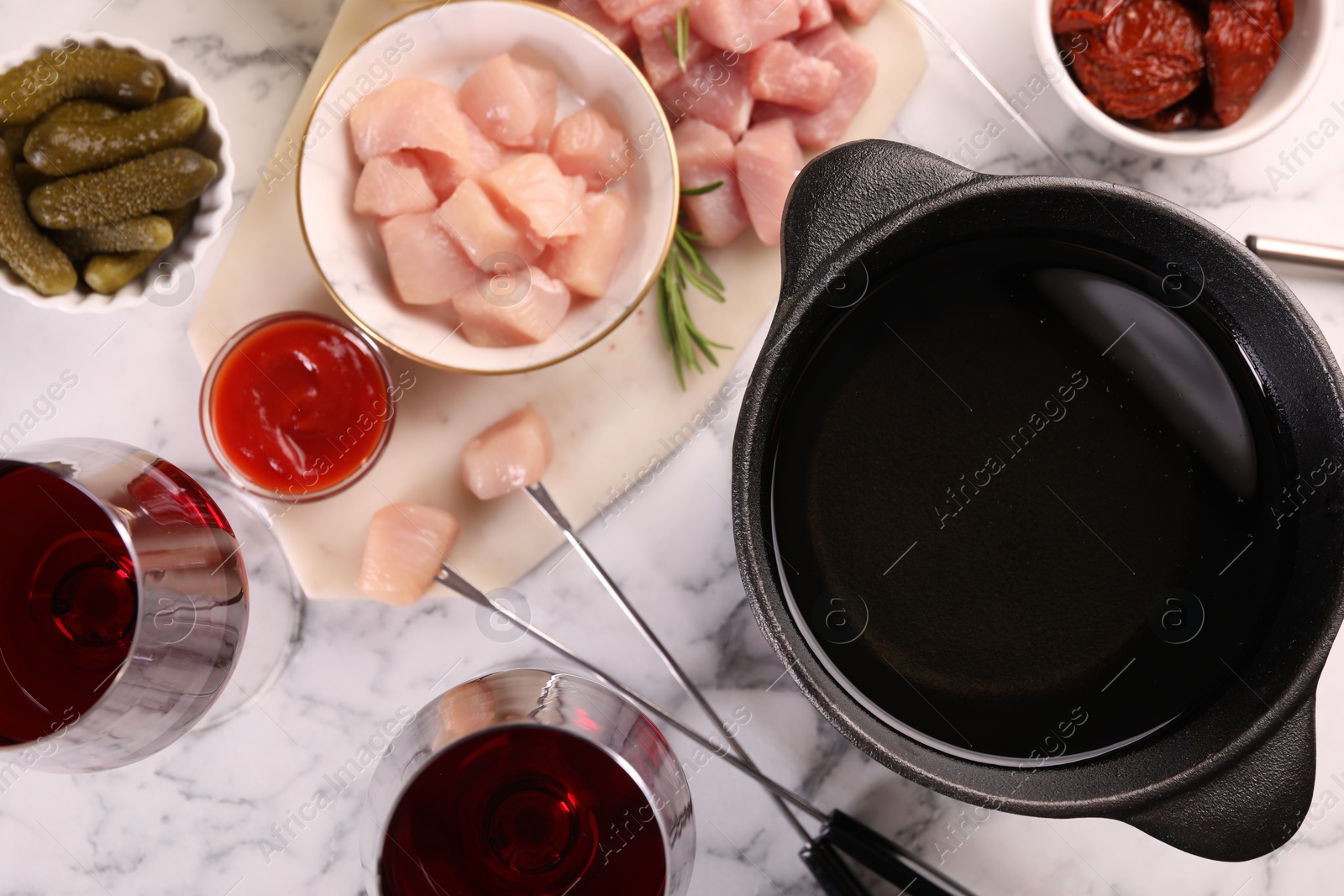 Photo of Fondue pot with oil, forks, raw meat pieces, red wine and other products on white marble table, flat lay