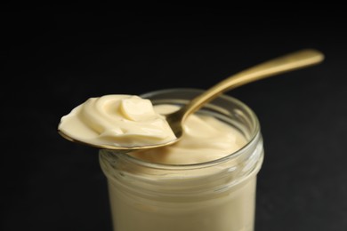 Photo of Jar of delicious mayonnaise and spoon on black table, closeup