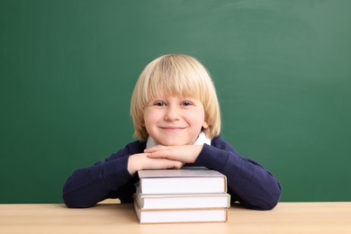 Happy little school child sitting at desk with books near chalkboard