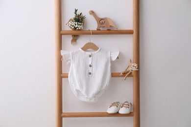 Photo of Baby bodysuit, shoes, toys and small bouquet on ladder near white wall