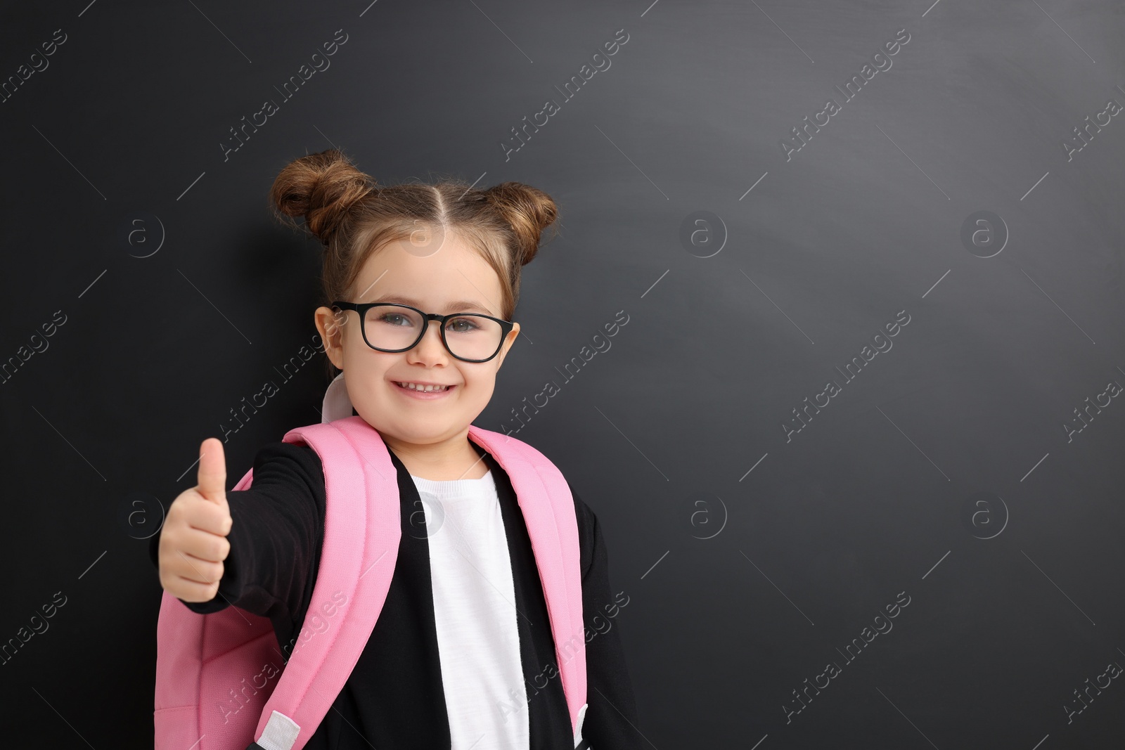 Photo of Happy little school child with backpack showing thumbs up near chalkboard. Space for text