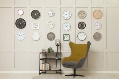 Armchair, console table and many different clocks hanging on white wall in room