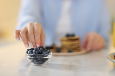 Photo of Woman taking blueberry from glass bowl at table, closeup