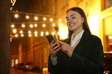 Smiling woman using smartphone on night city street. Space for text