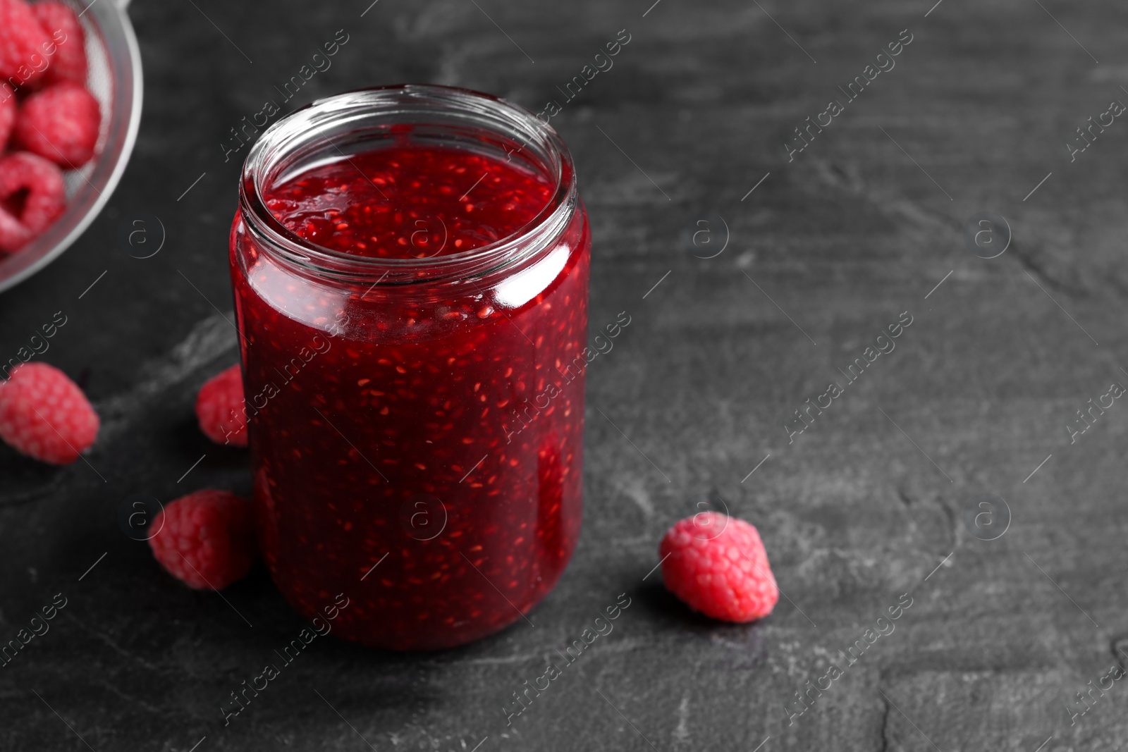 Photo of Delicious jam and fresh raspberries on black slate table, closeup. Space for text