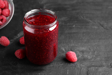 Photo of Delicious jam and fresh raspberries on black slate table, closeup. Space for text