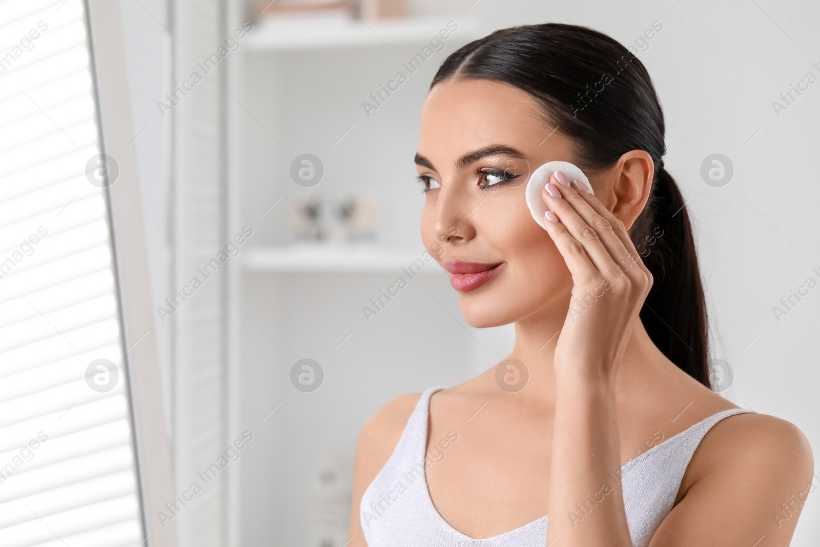 Photo of Beautiful woman removing makeup with cotton pad indoors, space for text