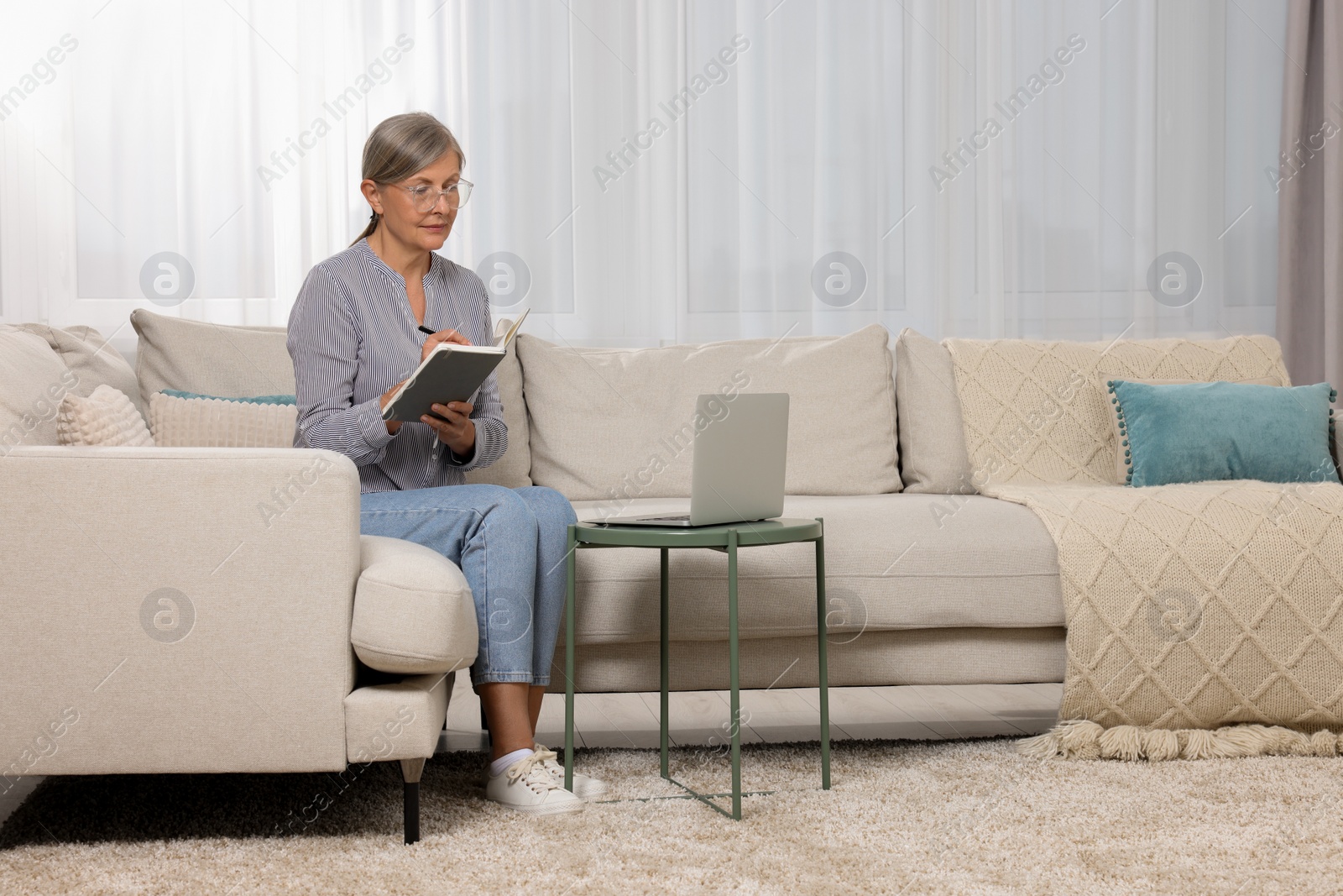 Photo of Beautiful senior woman writing something in notebook while working with laptop at home