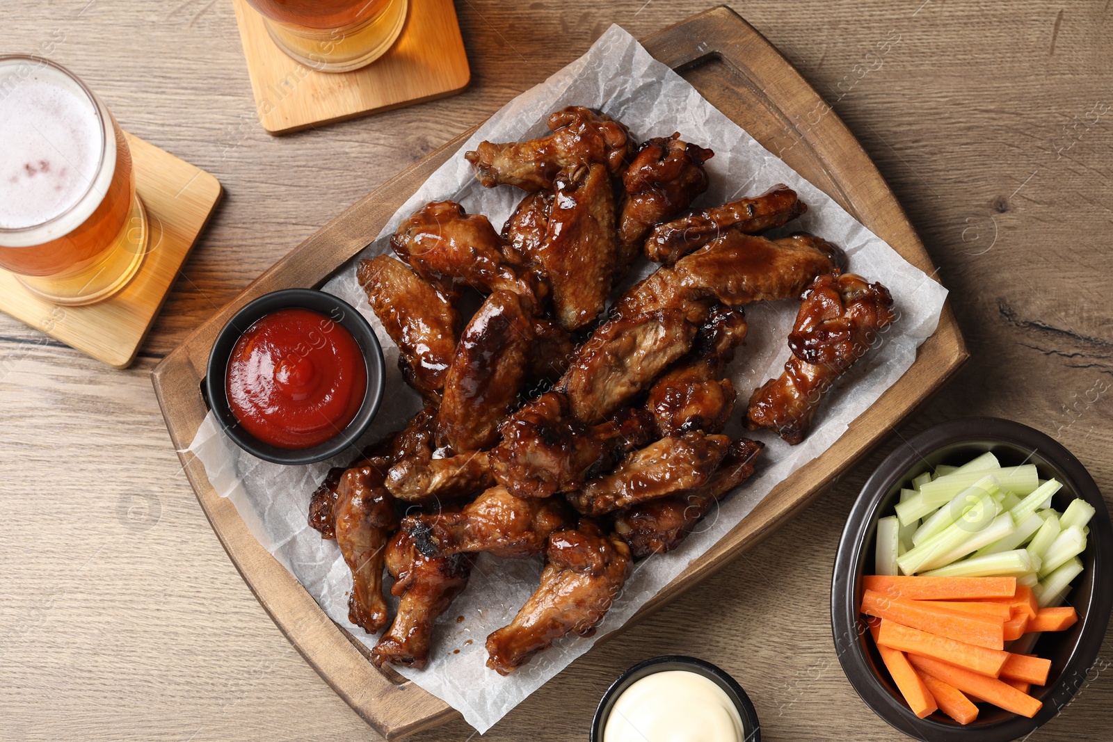 Photo of Tasty chicken wings, glasses of beer, sauces and vegetable sticks on wooden table, flat lay. Delicious snacks