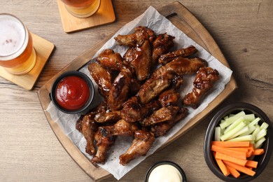 Photo of Tasty chicken wings, glasses of beer, sauces and vegetable sticks on wooden table, flat lay. Delicious snacks