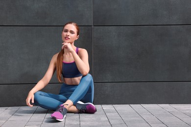 Photo of Beautiful woman in stylish gym clothes sitting near dark grey wall on street, space for text