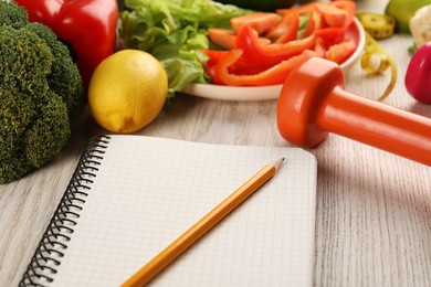 Photo of Healthy diet. Fresh vegetables, notebook, dumbbell and pencil on light wooden table, closeup