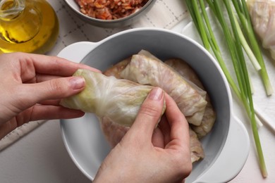 Woman putting uncooked stuffed cabbage roll into ceramic pot at white table, top view