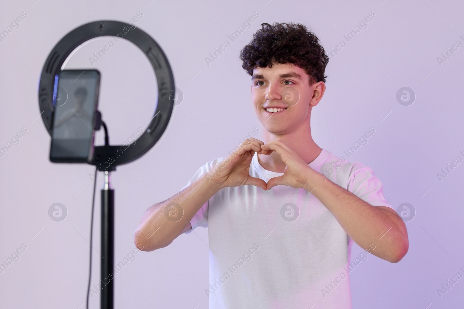 Photo of Smiling teenage blogger making heart gesture to his subscribers while streaming on white background