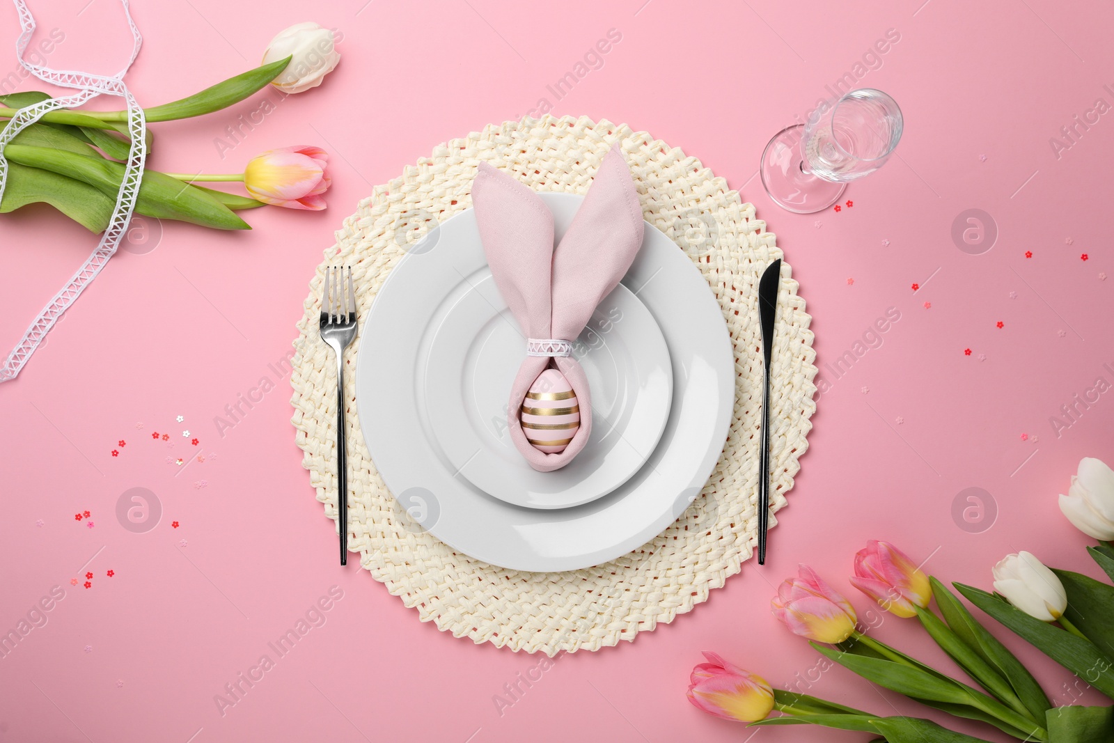 Photo of Festive table setting with painted egg, plates and tulips on pink background, flat lay. Easter celebration