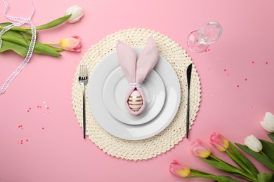 Photo of Festive table setting with painted egg, plates and tulips on pink background, flat lay. Easter celebration
