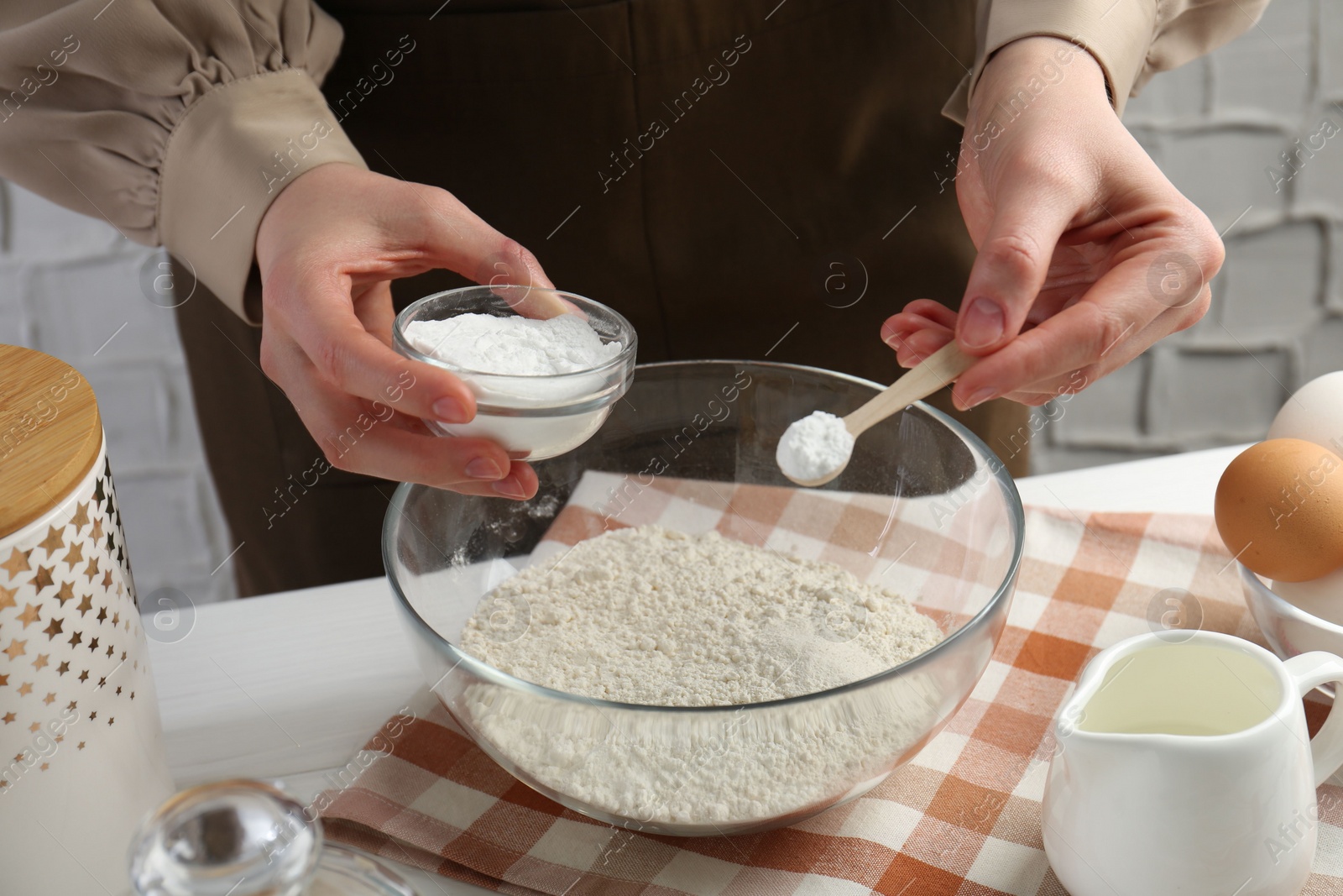 Photo of Woman with spoon and bowl of baking powder at white table, closeup