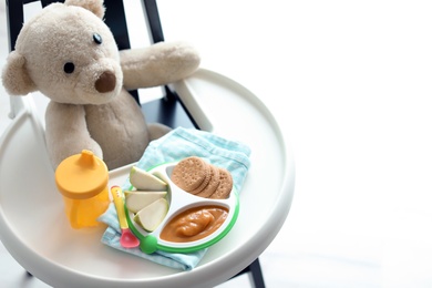 Photo of Plate with delicious baby food, drinking cup and teddy bear on highchair indoors