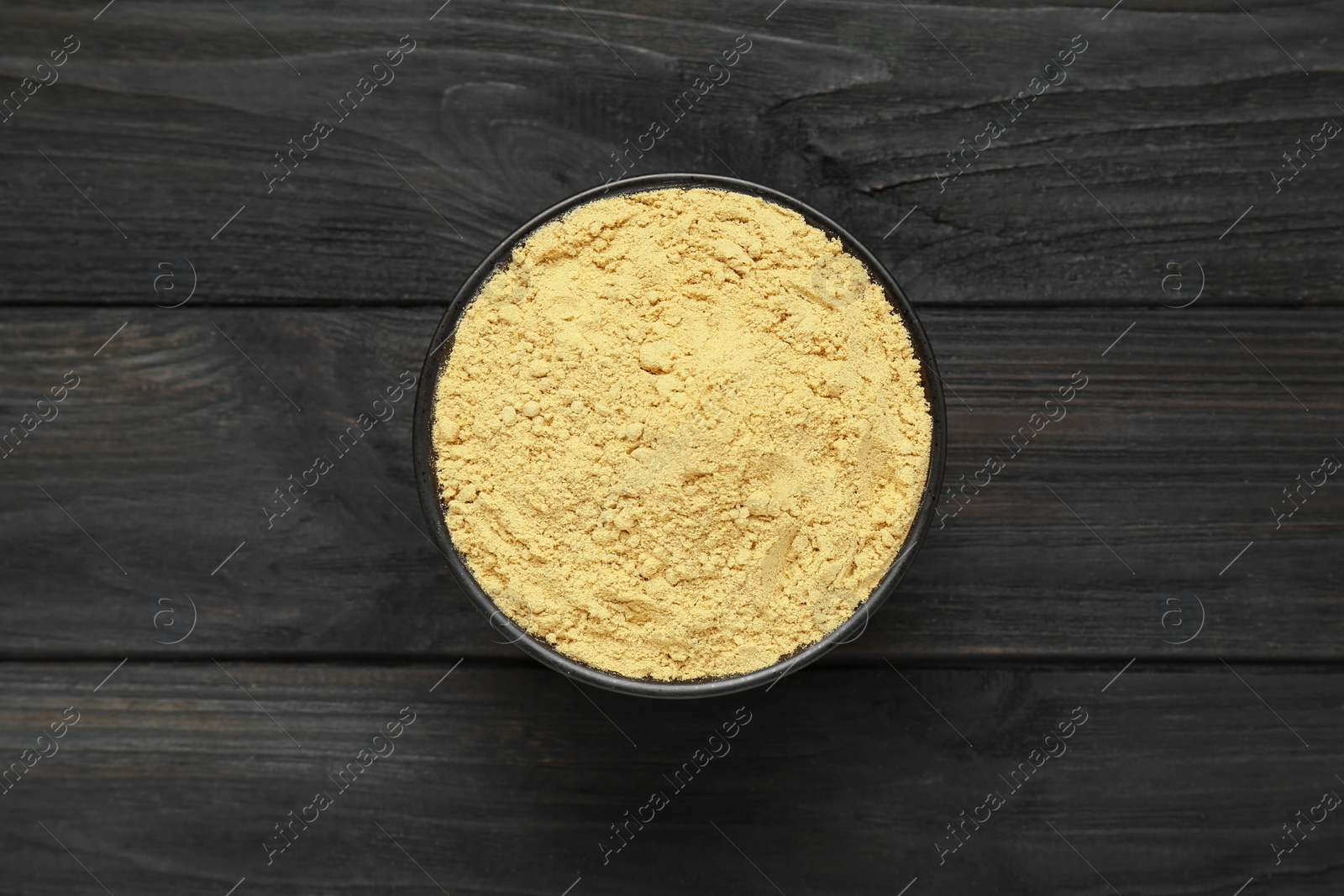 Photo of Bowl of aromatic mustard powder on black wooden table, top view