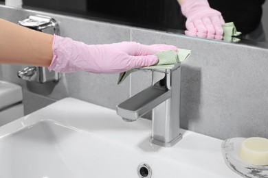Photo of Woman in glove cleaning faucet of bathroom sink with paper towel, closeup