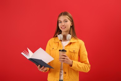 Teenage student with book, cup of coffee and headphones on red background