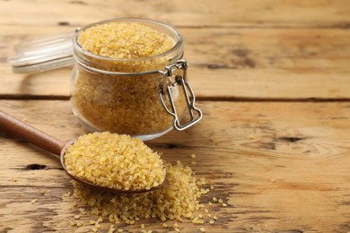 Glass jar and spoon with raw bulgur on wooden table, closeup. Space for text