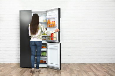 Photo of Young woman choosing food from refrigerator near white brick wall, space for text