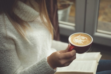 Photo of Woman with cup of coffee reading book near window indoors, closeup