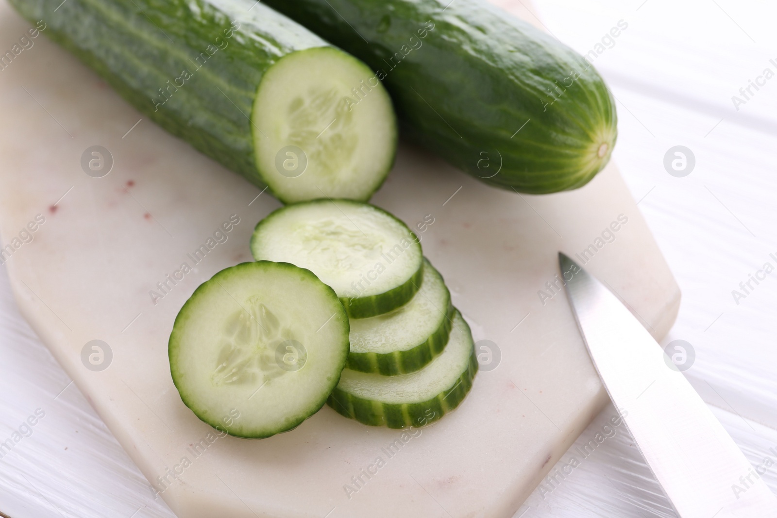 Photo of Cucumbers, knife and marble cutting board on white wooden table, closeup